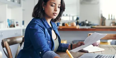 Een vrouw concentreert zich op papierwerk op een laptop in een zonnige kamer.