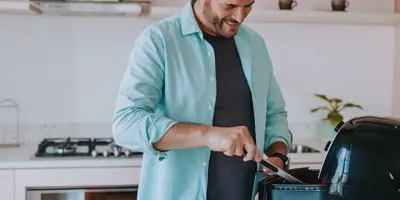 Een lachende man gebruikt een tang om de inhoud van een airfryer te roeren.