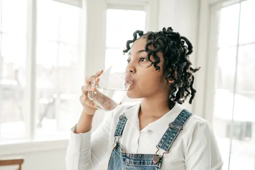 Een jong persoon staat in een helder verlichte kamer en drinkt een glas water.